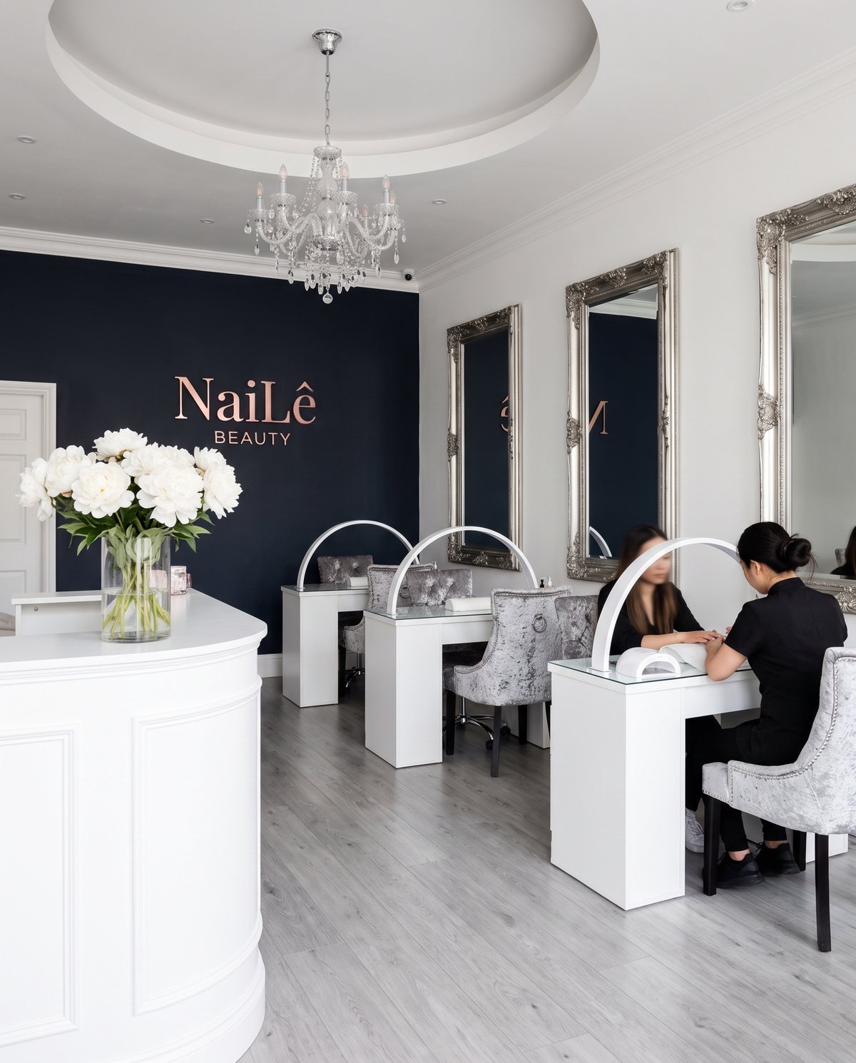 The salon: the reception desk, the sign wall, and desks under the silver mirrors with a client having her nails done